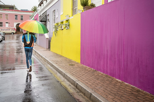 Rear view of a young man walking with a rainbow colored umbrella along a city street in the rain