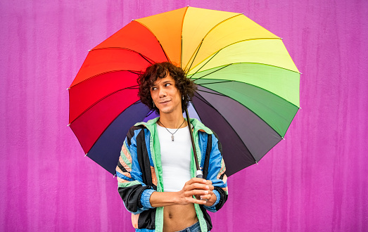 Young gay man holding a rainbow colored umbrella glancing sideways and smiling while standing in the rain in front of a pink wall