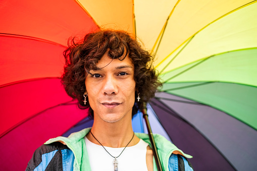 Portrait of smiling young gay man holding a rainbow colored umbrella while standing outside in the rain