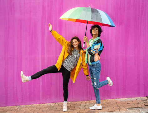 Portrait of two carefree young friends dancing with a rainbow colored umbrella along a city sidewalk in front of a pink wall