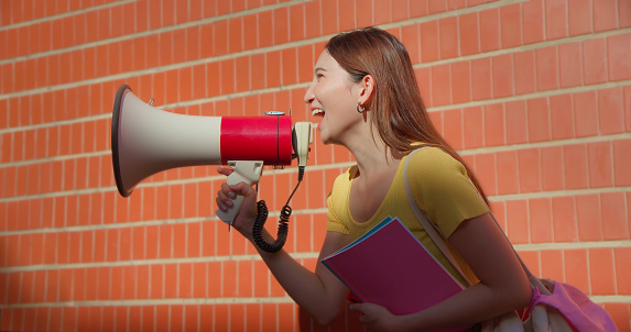 side view happy young attractive asian brunette long hair female student use megaphone happily in fornt of red brick wall