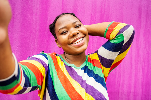 Smiling young African woman wearing a colorful striped sweater taking a selfie while standing in front of a pink wall outdoors