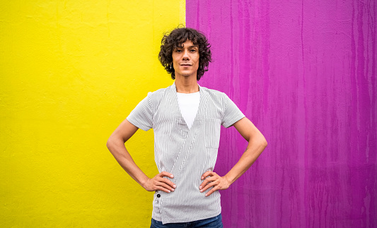 Portrait of a confident young gay man standing with his hands on his hips outside on a city sidewalk in front of a multi-colored wall