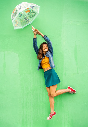 Portrait of a young woman smiling while holding onto an umbrella and floating in mid air against a green wall