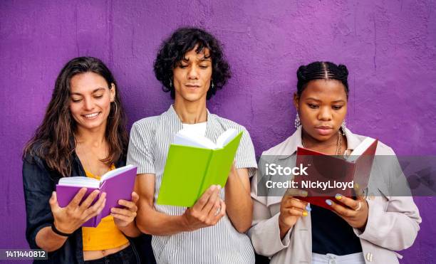 Three Young Friends Reading Books While Leaning Against A Purple Wall Stock Photo - Download Image Now