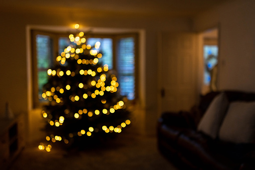 A Wide shot defocused Christmas tree with yellow lights inside a family home next to the sofa. It is low-key with minimal lighting.