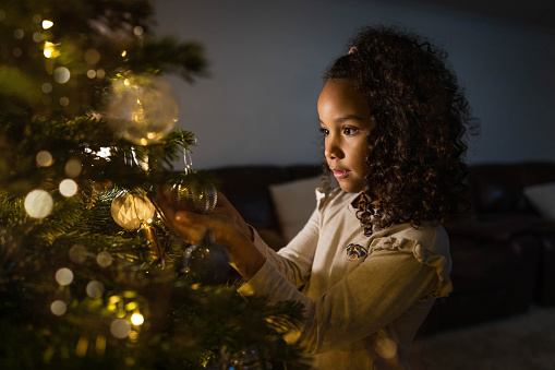 A young girl is holding up a bauble on a Christmas tree which has string lights on it illuminating her face. She is holding the bauble in amazement wearing pyjamas.