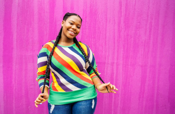 Smiling young woman with long braids standing against a pink wall Portrait of a smiling young African woman wearing a colorful striped sweater holding her braids while standing outside in front of a pink wall black woman hair extensions stock pictures, royalty-free photos & images