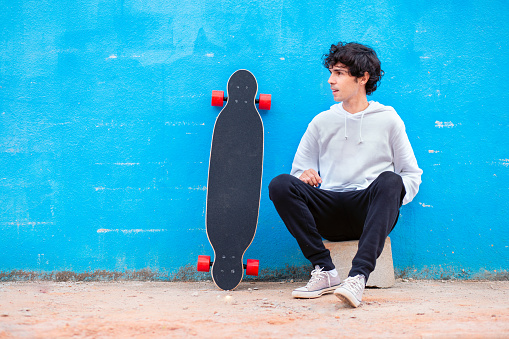 Portrait of a young man sitting with a skateboard at his side and blue wall in the background.