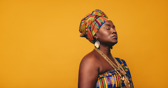 Ghanaian woman wearing traditional clothing against a yellow background. Mature black woman dressed in colourful Kente cloth and golden jewellery. Confident woman embracing her rich West African culture.