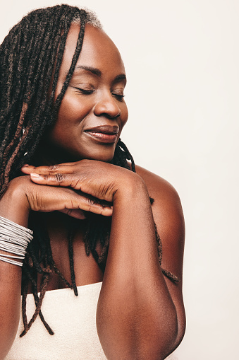 Elegant woman with dreadlocks wearing a bath towel and light make-up in a studio. Mature woman standing against a white background with her eyes closed. Self-confident woman pampering her ageing body.