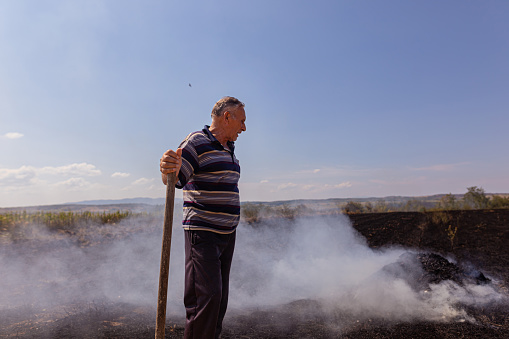 Side view of senior villager standing by the burnt and smoky fields, holding the shovel.