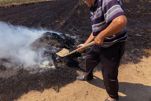 Side view of a senior farmer digging with a shovel burnt  and smoky ground in the field after a fire