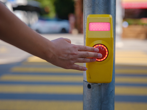 Close-up of a crosswalk signal button taken at a pedestrian controlled crossing. Hand pushing button to cross