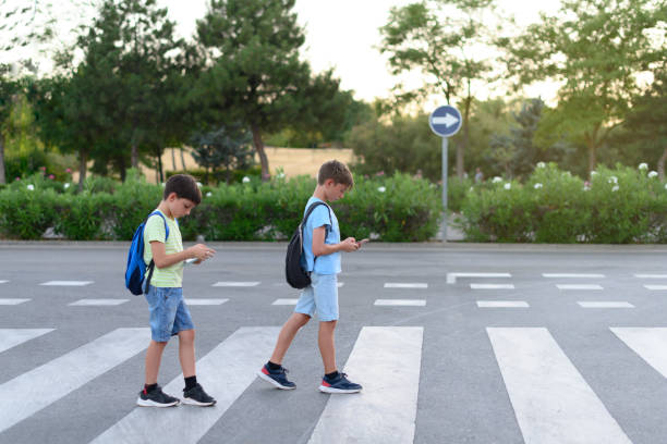 deux enfants regardent leur téléphone portable en traversant un passage pour piétons dans la rue. concept de dépendance au téléphone mobile - digital native photos et images de collection