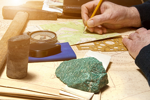 Geologist works with maps. On his desk are: map case, geological hammer, compass, magnifying glass, drill core, rock samples, topographic and geological maps