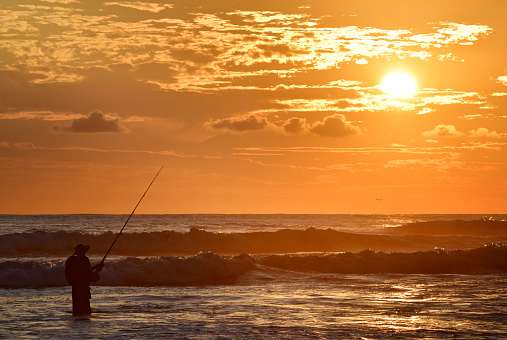 This photograph displays a silhouette of a fisherman in the ocean at sunset.  The sun is setting, the sky is orange and there are a few clouds.