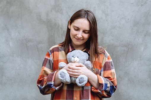 Girl holding a cute teddy bear toy in her hands. Woman in a plaid shirt on a background of gray concrete wall. The brunette looked down.