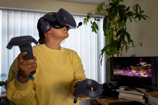 A front-view waist-up shot of a young woman playing a video game using a virtual reality headset, she is holding joystick controllers in each hand and she is having fun at her home in Northeastern England.