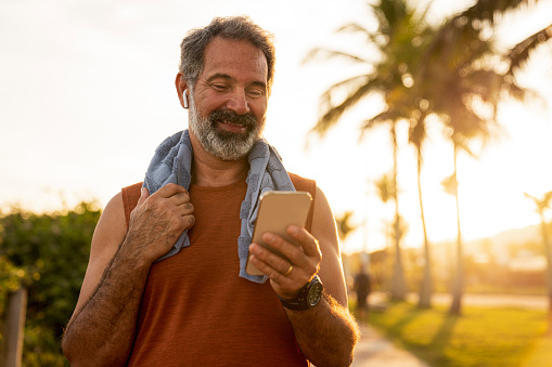 A motivated senior athlete, standing with a smartphone in his hand and a towel around his neck. He catches his breath after running and rests