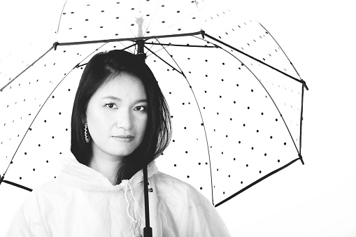 Studio shot of beautiful Asian woman with long black hairs wearing raincoat and hiding under transparent with black dots umbrella with copy space in white background. Black and white image