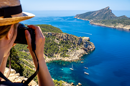 Rear view of a young white woman with a camera and hat above the cozy bay Cala en Basset nearby the village Sant Elm at the most west side of Mallorca with anchored boats and the island Sa Dragonera.