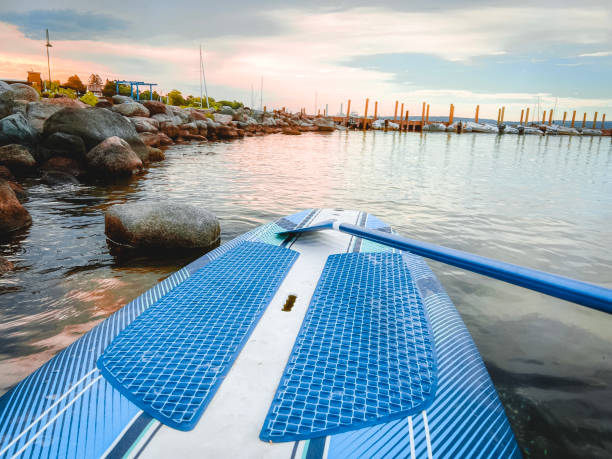 A Stand-Up Paddle Board and Oar Resting on the Shore at dawn. Located within a bay of water with views of a pier with moored sailboats and the coastline. Dawn or dusk dramatic landscape scene.