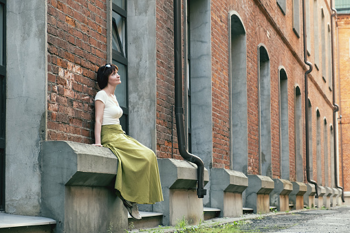 Woman in a long skirt sits on the basement cornice of an old building with large windows and a red brick facade. Woman looks up thoughtfully. Walking through historical places, travel.