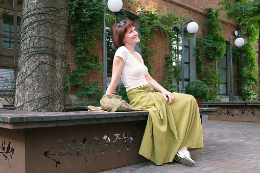 Woman with dark red hair sitting on a bench and smiling. Walks around the city. Middle-aged woman waiting for a date.