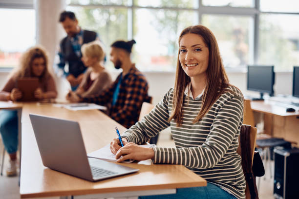 istock Happy mid adult student using laptop while writing notes in the classroom and looking at camera. 1411477884