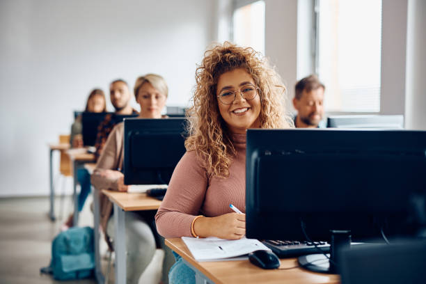 Happy college student taking notes on computer class and looking at camera. Young happy woman writing notes while using desktop PC in the classroom and looking at camera. computer lab stock pictures, royalty-free photos & images