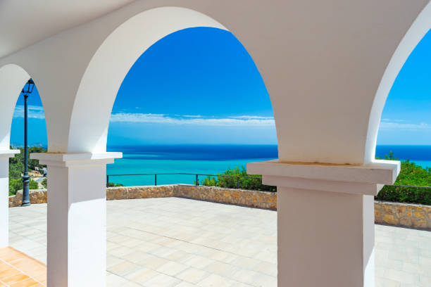 Blonde woman in a pink dress stands on a balcony, gazing out at the serene view of green trees and the tranquil sea beyond, enjoying a peaceful moment of contemplation