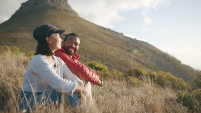 Couple sitting and watching the sunset on a mountain. Happy man and woman enjoying their vacation destination. Girlfriend and boyfriend in love in nature after hiking. Married couple relaxed outdoors