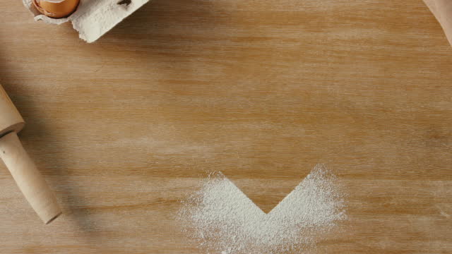 Stop motion of a heart and baking supplies from above. Overhead of heart shape image being made with flour on a wooden kitchen counter with cooking tools. Oven mitts, rolling pin, and eggs on a table