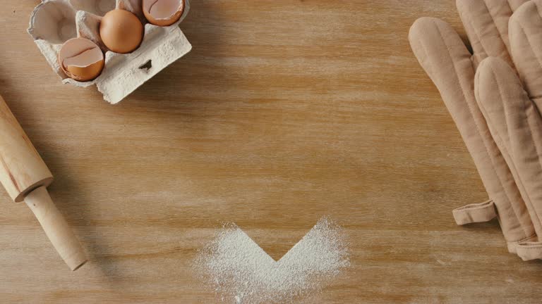 Stop motion of a heart and baking supplies from above. Overhead of heart shape image being made with flour on a wooden kitchen counter with cooking tools. Oven mitts, rolling pin, and eggs on a table