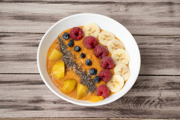 Orange smoothie in a white bowl with raspberries, carrots, mango, banana, chia seeds on a wooden background.