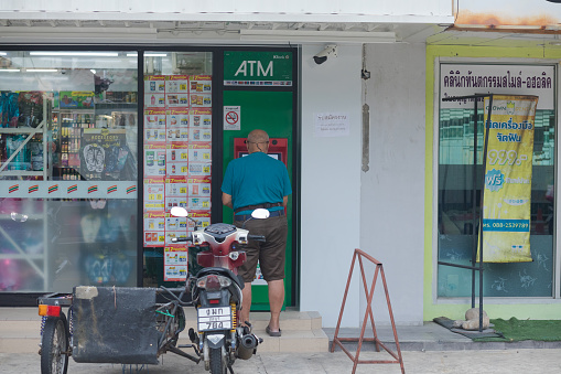 Rear view of bald headed thai man at ATM. Man is wearing casual clothing and is using ATM outsode of a small supermarket. In foreground a motorcycle with side carrier is standing. Scene is in Bangkok Chatuchak
