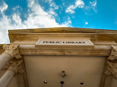 Low angle view of a stone portico with chandelier hanging. Looking up at the words Public Library engraved into the stone. Set against a perfect blue sky.