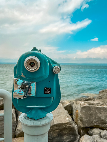 Teal colored vintage style viewfinder telescope positioned in the forefront of view. Telescope on the shoreline looks out over the beautiful turquoise blue lake waters. Water meets a cloudy blue daytime sky at the horizon. Located at Lake Michigan in Petoskey, Michigan, USA.