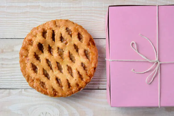 Top view of a fresh baked apple pie on a rustic white wood table next to a pink bakery box tied with string.
