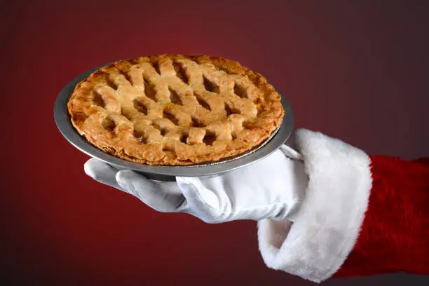 Santa Claus hand holding a fresh baked apple pie over a light to dark red background. Horizontal format showing only Santa's hand and arm.