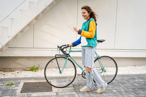One woman young adult caucasian female walking by the university building holding bicycle and digital tablet in day real people copy space happy smile joyful full length side view