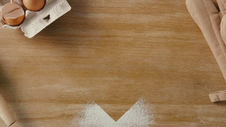 Closeup of a Heart  shape flour image and rolling pin , baking mitts and a small tray of eggs on color wooden table in a kitchen.