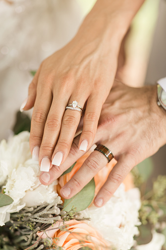 Manos de una pareja de novios con sus anillos de boda sobre un suave ramo de boda en pastel con rosas de color melocotón, peonías blancas y eucalipto verde salvia en Palm Beach, Florida photo