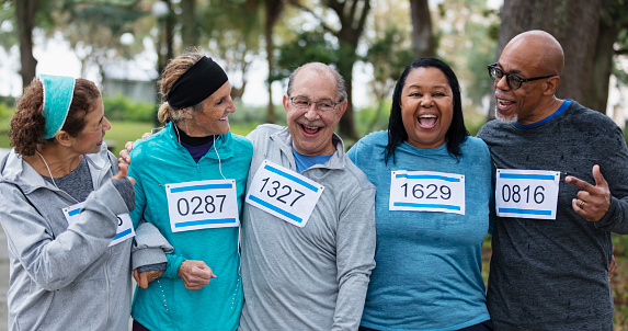 A multiracial group of five friends in their 50s and 60s standing side by side ready to run in a race, wearing numbered sports bibs on their shirts. The African-American woman is looking toward the camera.