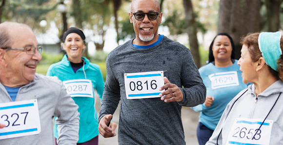 A multiracial group of five men and women in their 50s and 60s running in a race at a park, wearing marathon bibs. The focus is on the African-American man, a senior in his 60s.