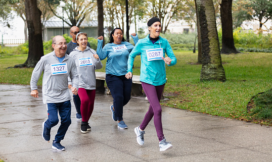 A multiracial group of five adults running a race together in a park. They are wearing numbered sports bibs. The focus is on the mature African-American woman, in her 50s, and her friend running beside her, a senior Hispanic woman in her 60s.