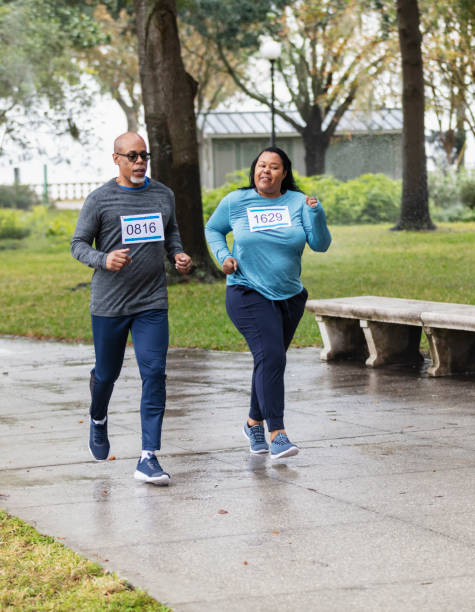 African-American couple running in race together An African-American couple running in a race together, side by side, through a park, wearing marathon bibs. The senior man is in his 60s and his partner is in her 50s. body positive couple stock pictures, royalty-free photos & images