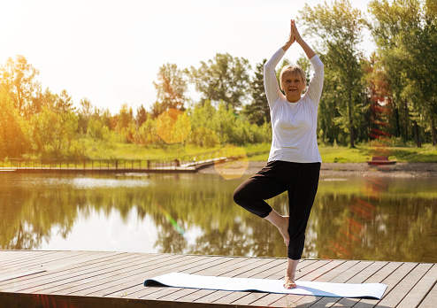 Full-length portrait of a Caucasian elderly woman in yoga vrikshasana poses, tree pose, against the background of a lake in a park. Health and active lifestyle of the elderly