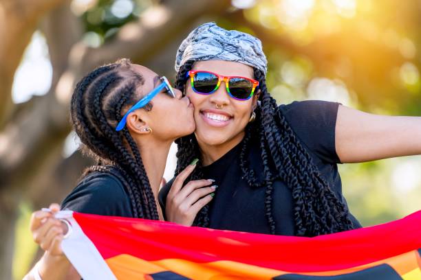 une jeune lesbienne afro-latine embrassant sa femme tenant une bannière arc-en-ciel avec le mot proud écrit dessus - couple black american culture kissing photos et images de collection